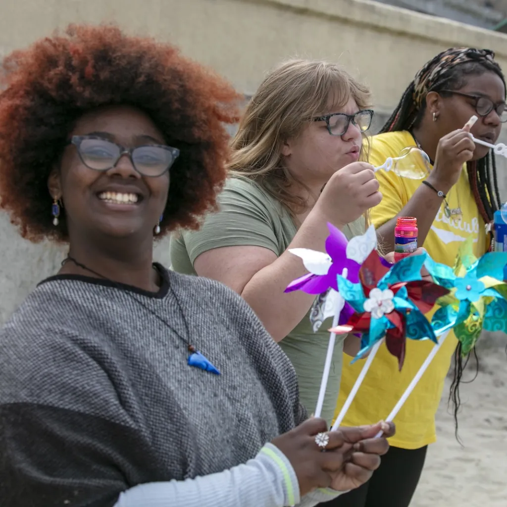 Four women smile and laugh while blowing bubbles and holding colorful pinwheels on a sandy surface with a concrete wall in the background.