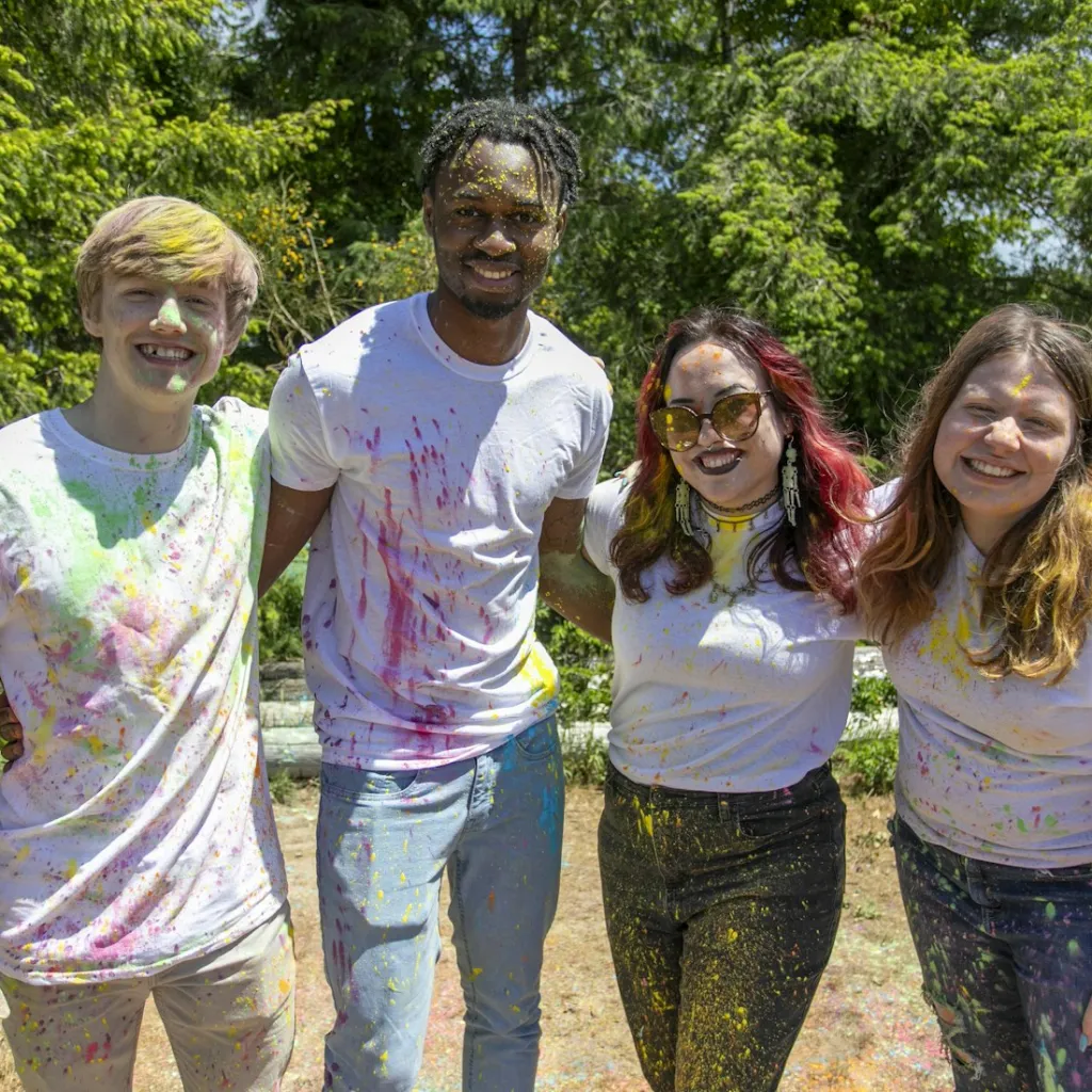 A group of four young adults wearing white t-shirts, covered in colorful powder, stand together outdoors smiling, with trees and greenery in the background.