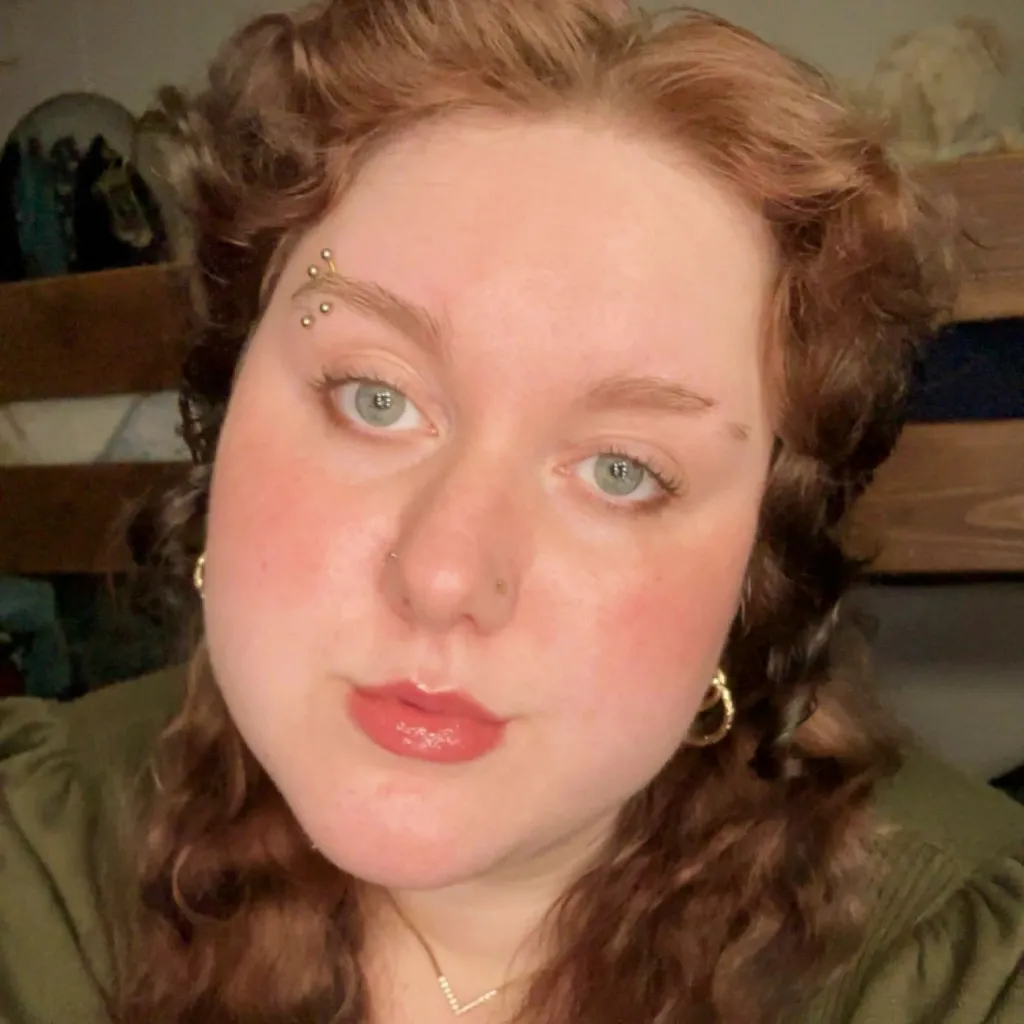Headshot of Brina Williams wearing an olive green top and gold hoop earrings, with curly auburn hair, multiple eyebrow piercings, a nose ring, and red lipstick, in an indoor setting.