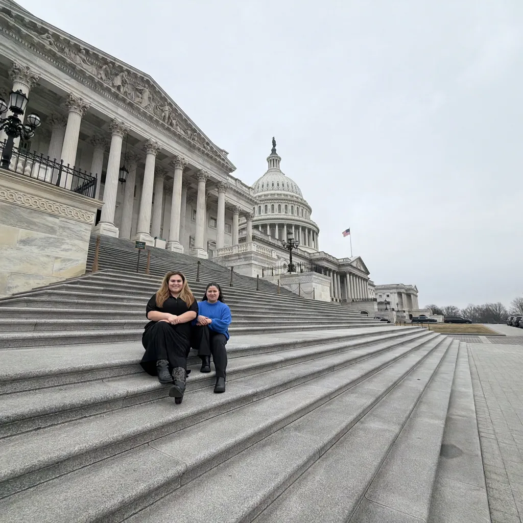 Two young people sitting on steps of US Capital building. One wearing black outfit and one wearing blue sweater 