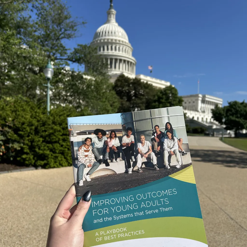 A hand holding a copy of "Improving Outcomes for Young Adults and the Systems that Serve Them: A Playbook of Best Practices" in front of the U.S. Capitol building on a sunny day.
