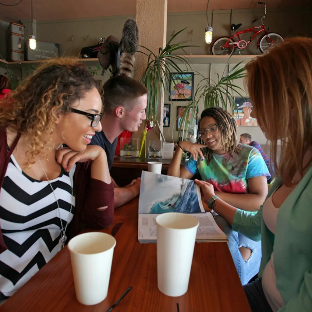 Four people sit around a wooden table in a café, engaged in conversation. One person holds open a magazine showing a large wave, while the others smile and lean in. The background features plants, artwork, and a red bicycle on the wall.
