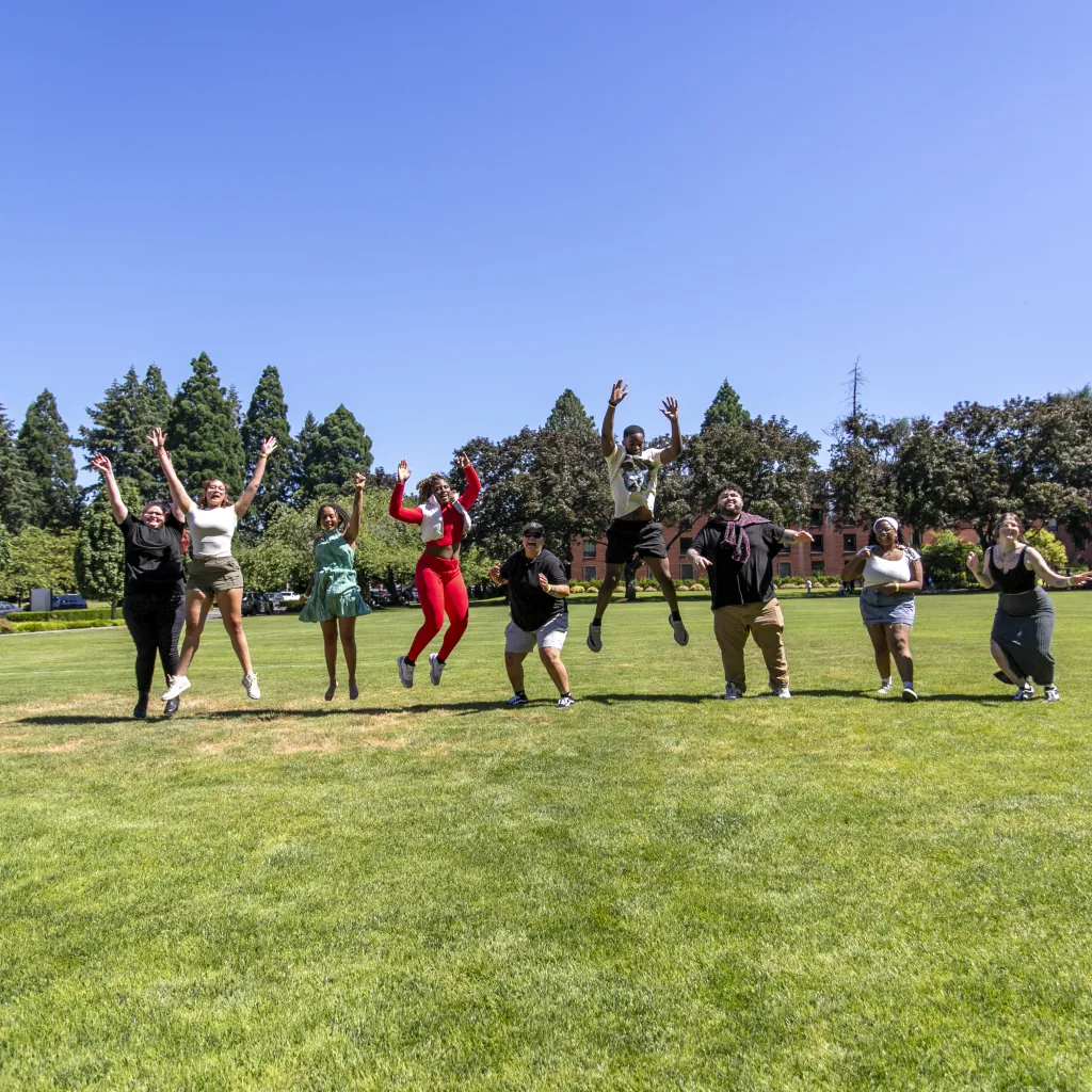 group of young people jumping to celebrate on large grassy area with trees and blue sky behind them