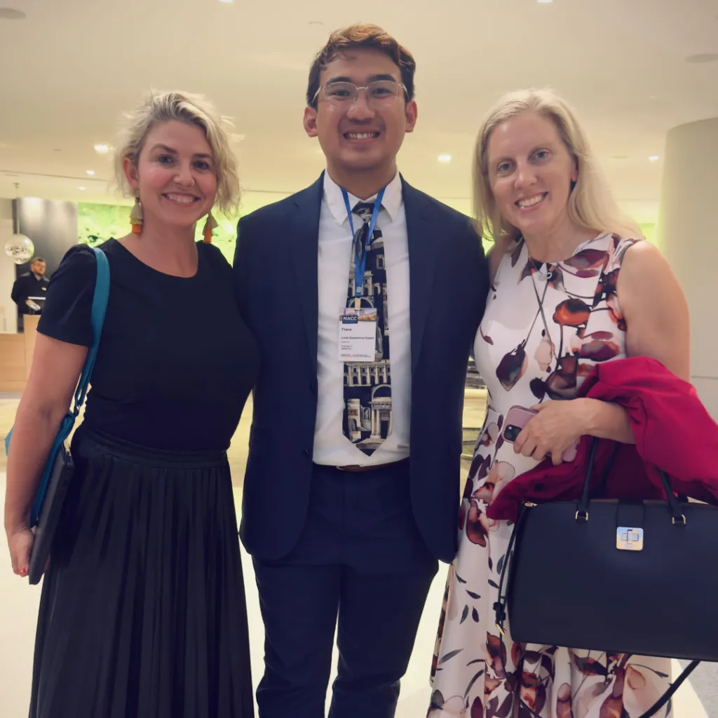 Three people smiling and standing together indoors. The person in the center is wearing a suit and tie with a name badge, while the two women on either side are dressed in business-casual attire, one in a black skirt and the other in a floral dress.