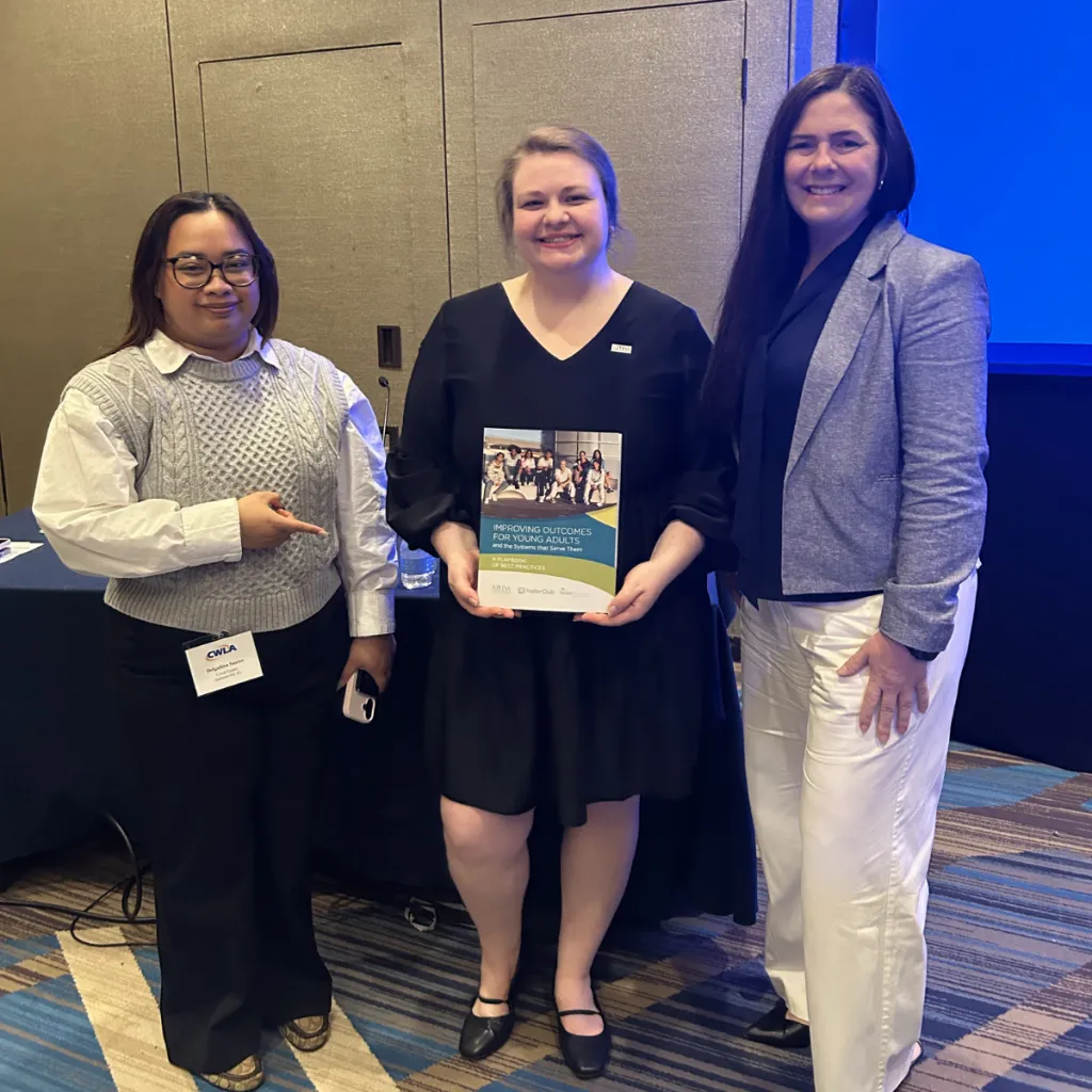 Three people stand together smiling at what appears to be a professional conference. The person on the left wears glasses, a cable-knit sweater, and a CWLA (Child Welfare League of America) name badge. The woman in the center holds a publication titled "Improving Outcomes for Young Adults and the Systems that Serve Them." The woman on the right is wearing a grey blazer and white pants. The setting is a hotel meeting room with blue accent lighting in the background.