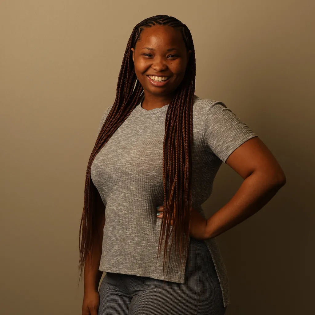 Headshot of Dejah Carlock smiling with one hand on her hip, wearing a grey t-shirt, with long braids, against a tan background.