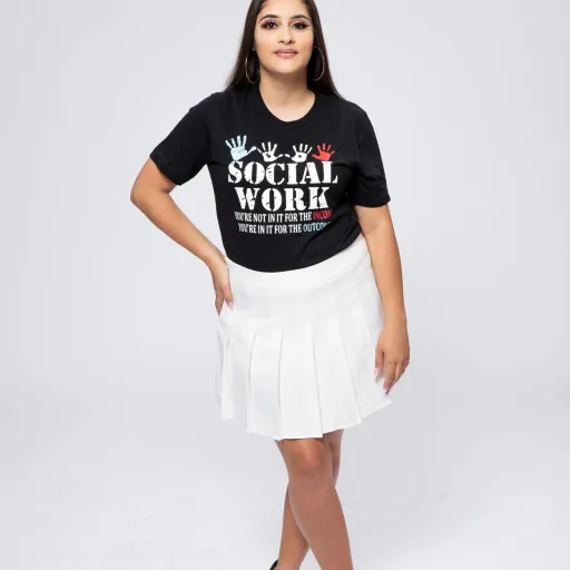 Mayda Berrios stands against white background with black t-shirt that says "Social Work - You're not in it for the income. You're in it for the outcome." 4 handprints are on the t-shirt as well.