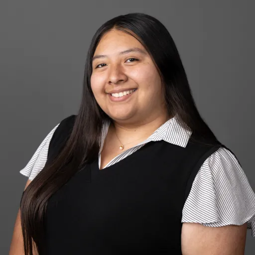 Headshot of Clarissa Pena smiling, wearing a black vest over a striped collared shirt with a gold necklace, with long dark hair, against a grey background.