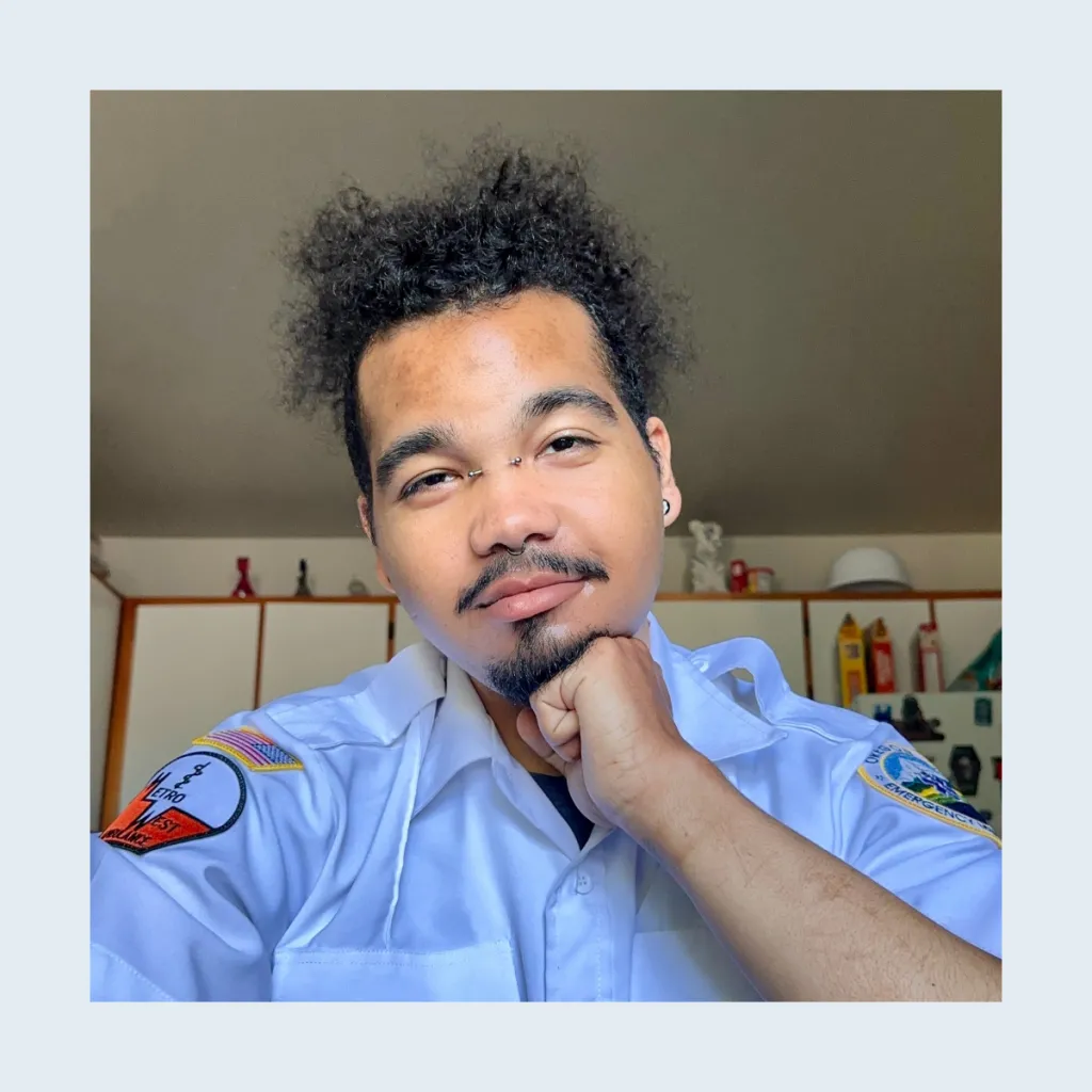 Headshot of Alexander Robbinson resting their chin on their hand, wearing a light blue uniform shirt with emergency services patches, with curly hair and a nose ring, in a home setting.