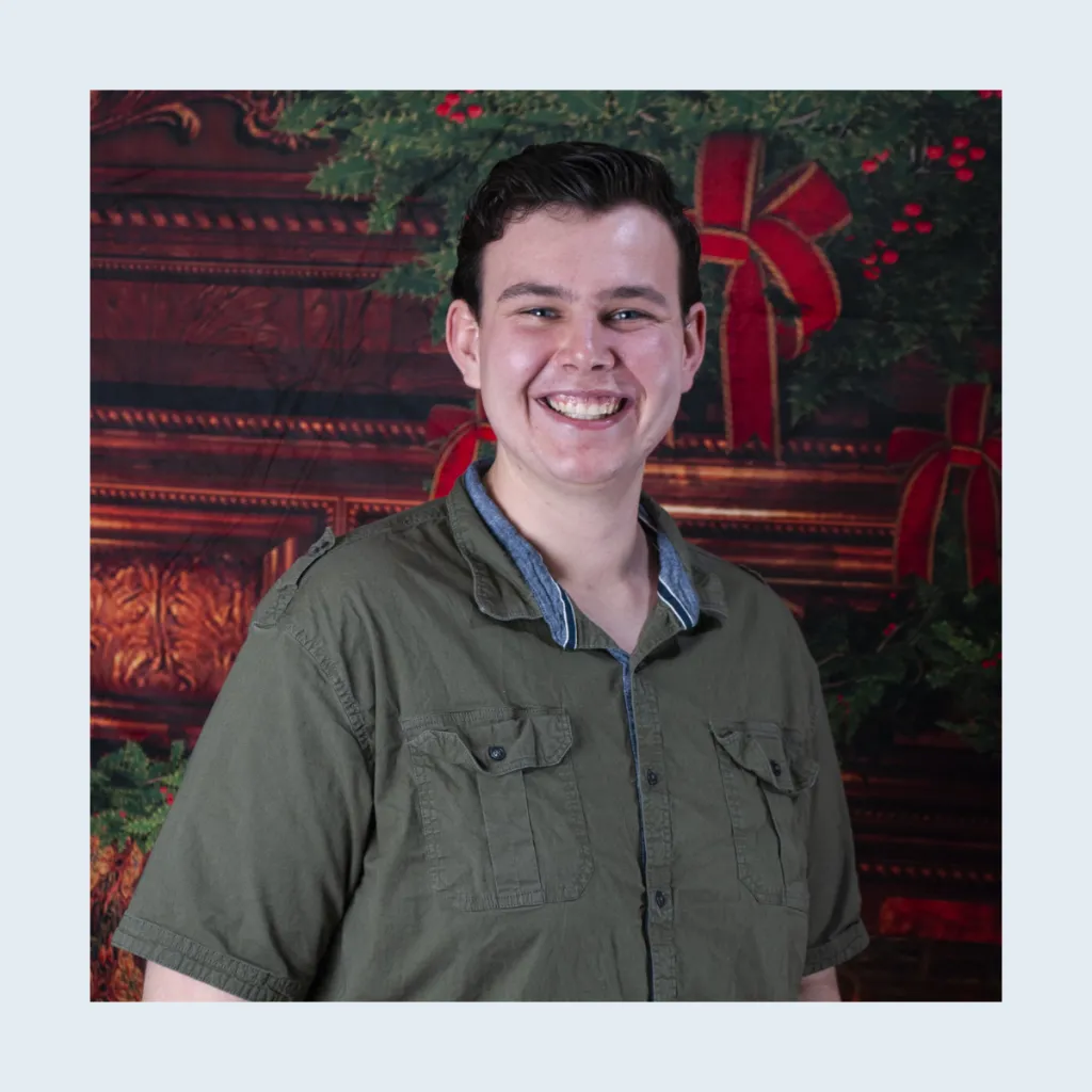 Headshot of Ash Fayal smiling, wearing an olive green button-up shirt, against a festive holiday backdrop with wreaths and red ribbons.