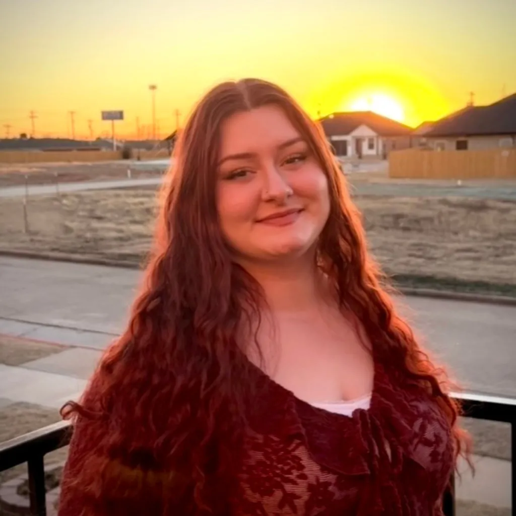 Headshot of Michalann Clark smiling, wearing a dark red lace blouse, with long wavy auburn hair, against a golden sunset backdrop.