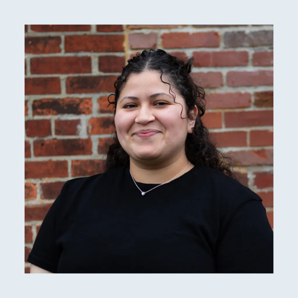 Headshot of Cinthia Garcia smiling, wearing a black t-shirt and a delicate silver necklace, with curly hair half pulled back, against a brick wall.