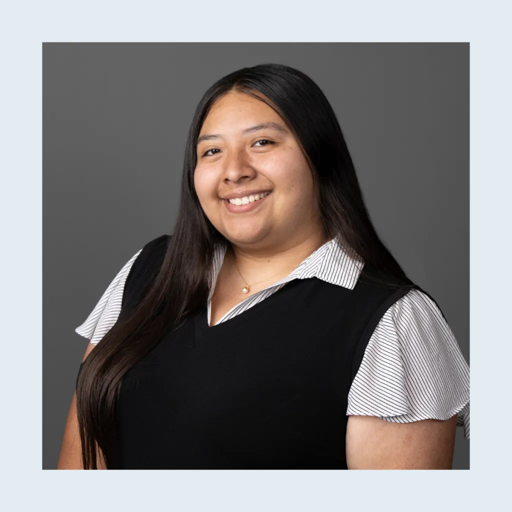 Headshot of Clarissa Pena smiling, wearing a black vest over a striped collared shirt with a gold necklace, with long dark hair, against a grey background.