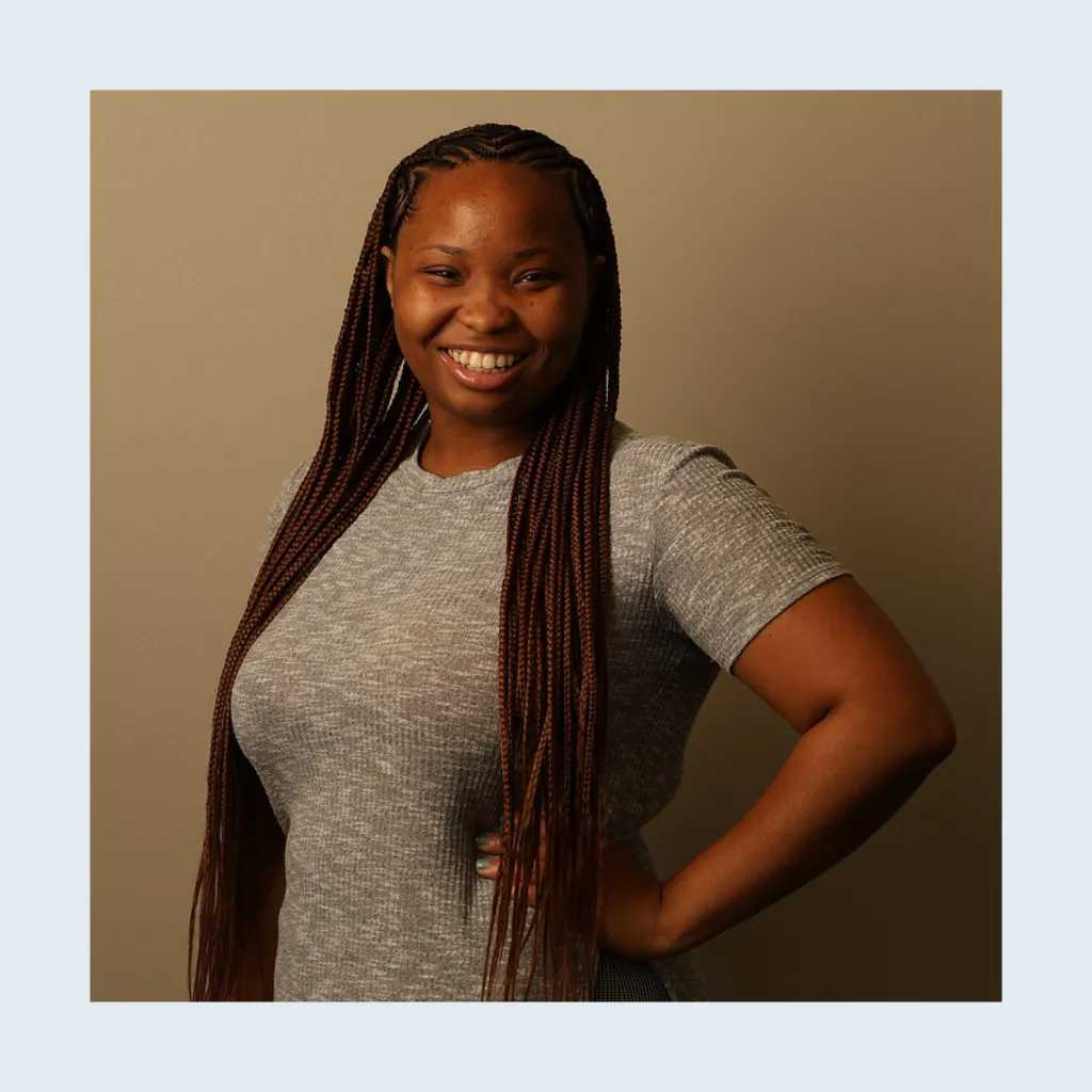 Headshot of Dejah Carlock smiling with one hand on her hip, wearing a grey t-shirt, with long braids, against a tan background.