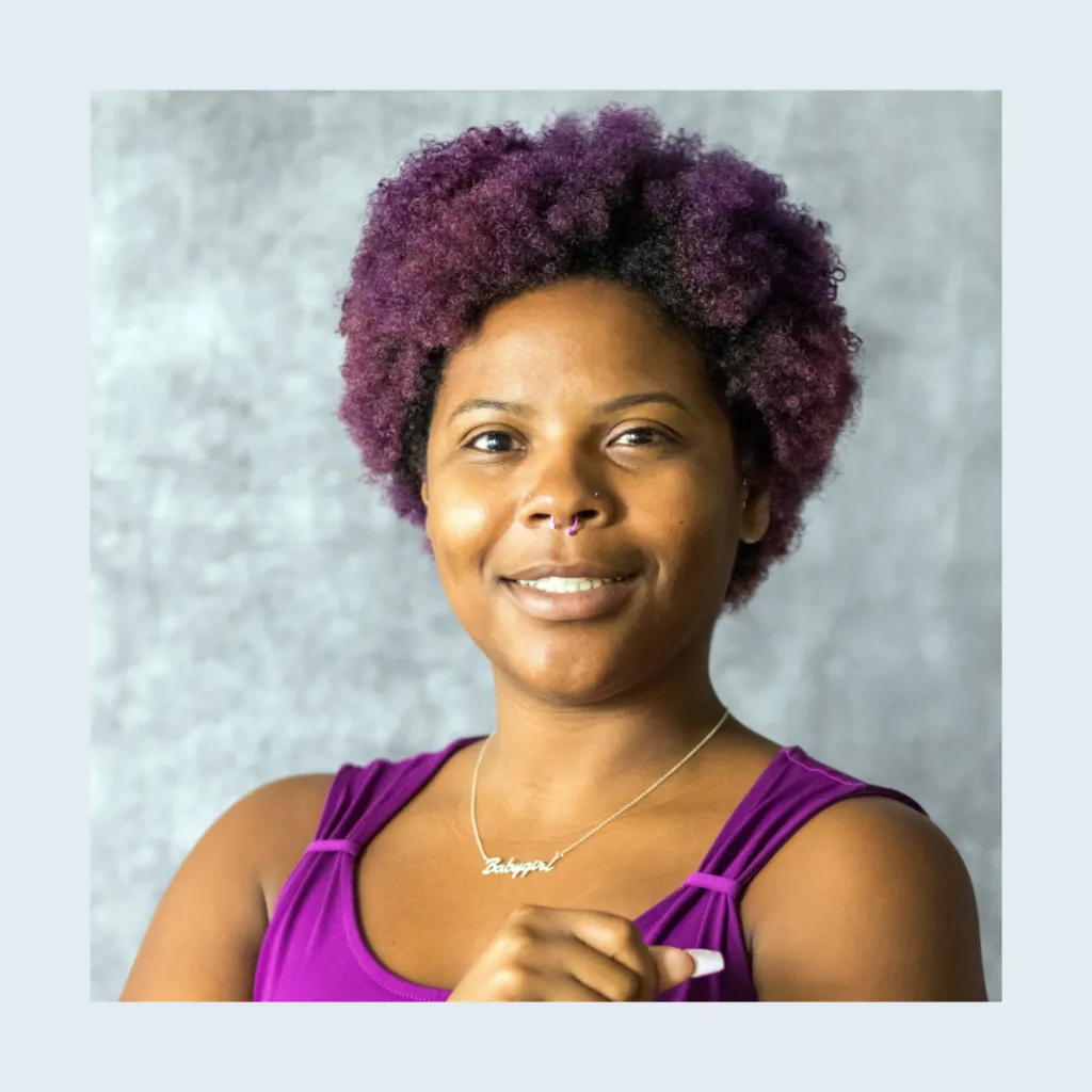 Headshot of Jasmine Solomon smiling, wearing a purple top and a nameplate necklace, with a purple natural afro and a nose ring, against a grey background.