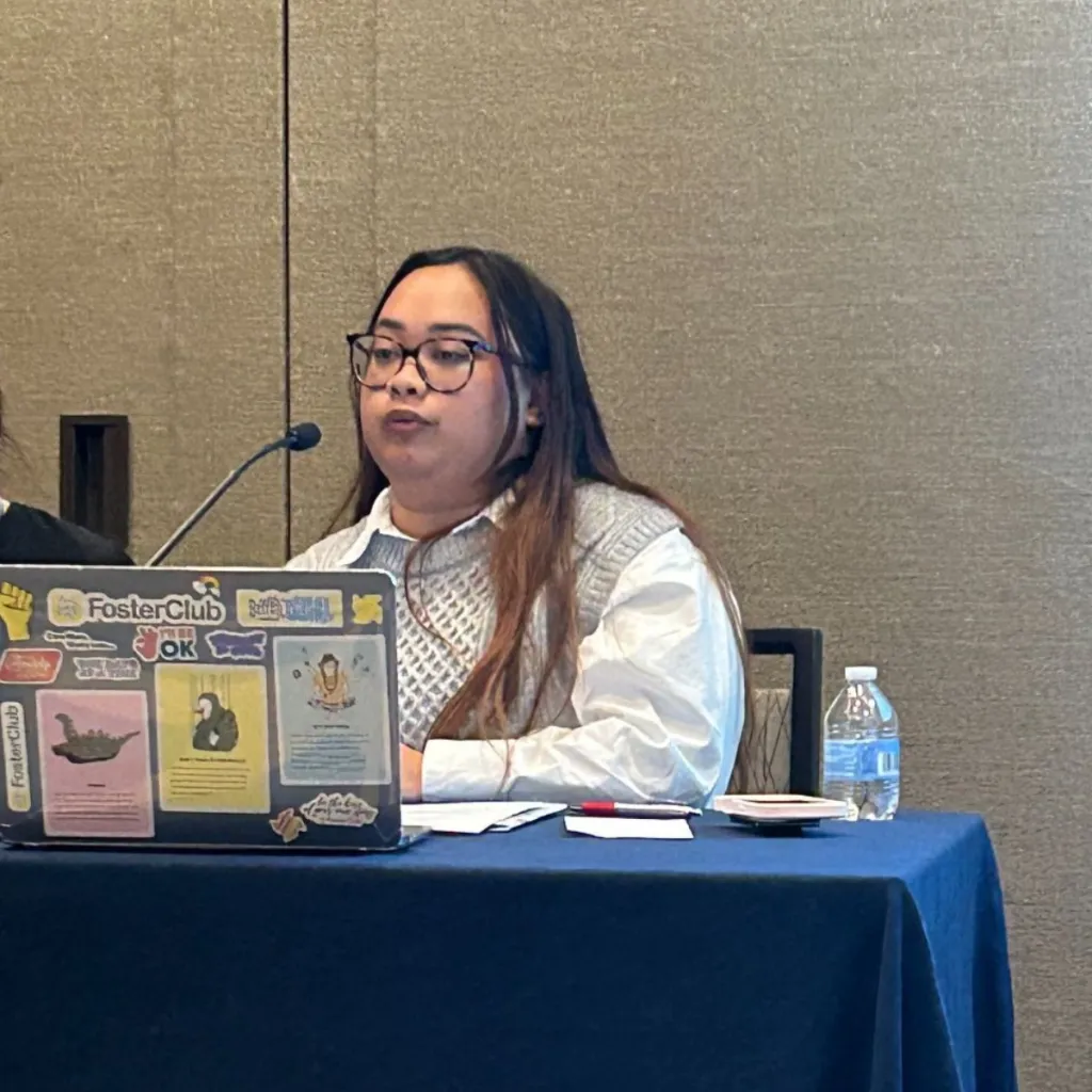 LEx Leader Dina speaks at a conference table microphone, wearing glasses and a grey sweater vest over a white collared shirt, with a sticker-covered FosterClub laptop open in front of them.