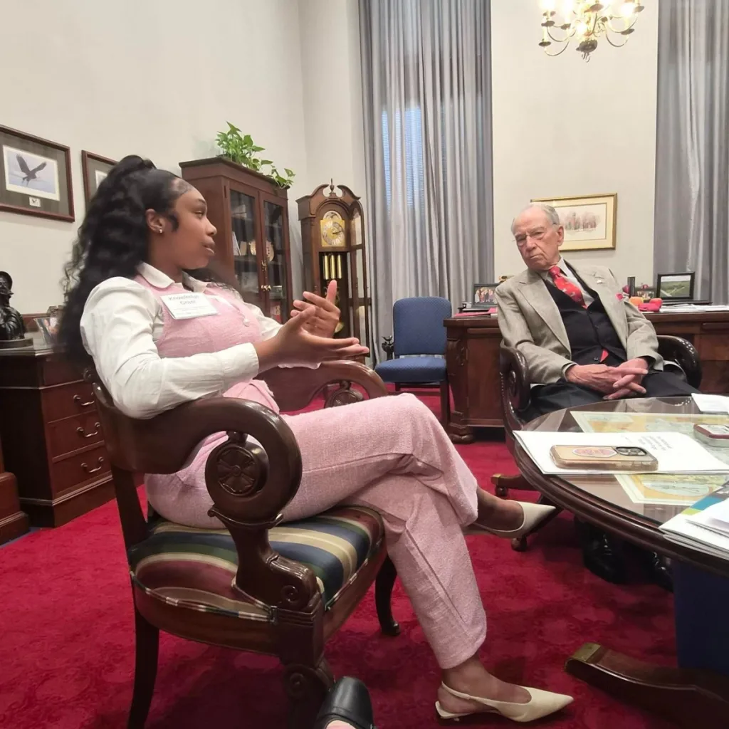 LEx Leader Knowledge speaks with a person in a formal Congressional office, wearing a pink outfit and name badge, gesturing while seated in an ornate wooden chair across from her listener, in a room with red carpet and dark wood furniture.