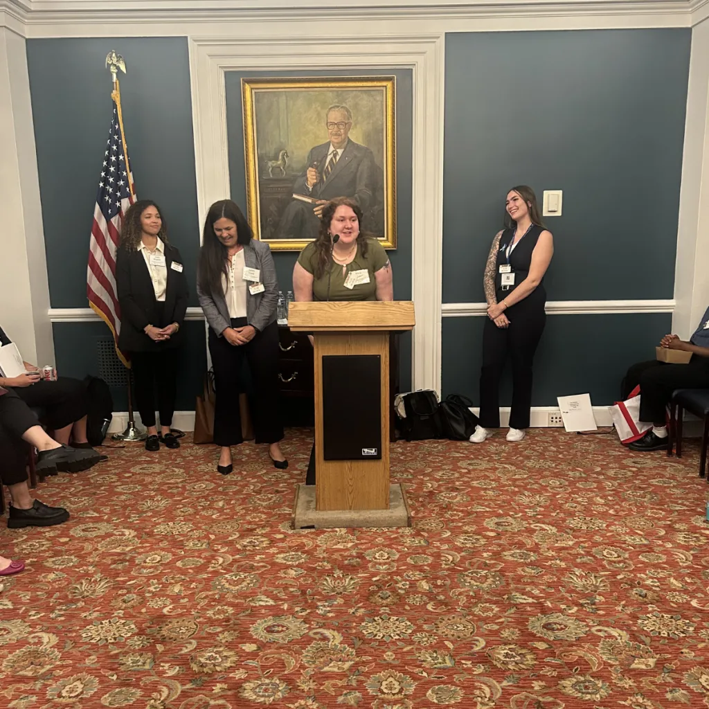 FosterClub LEx Leader Brina Williams speaks at a podium in a formal Congressional meeting room, flanked by FosterClub LEx Leader Natalie Clark, one partner staff, and a state leader, with an American flag and a portrait painting on the teal wall behind them