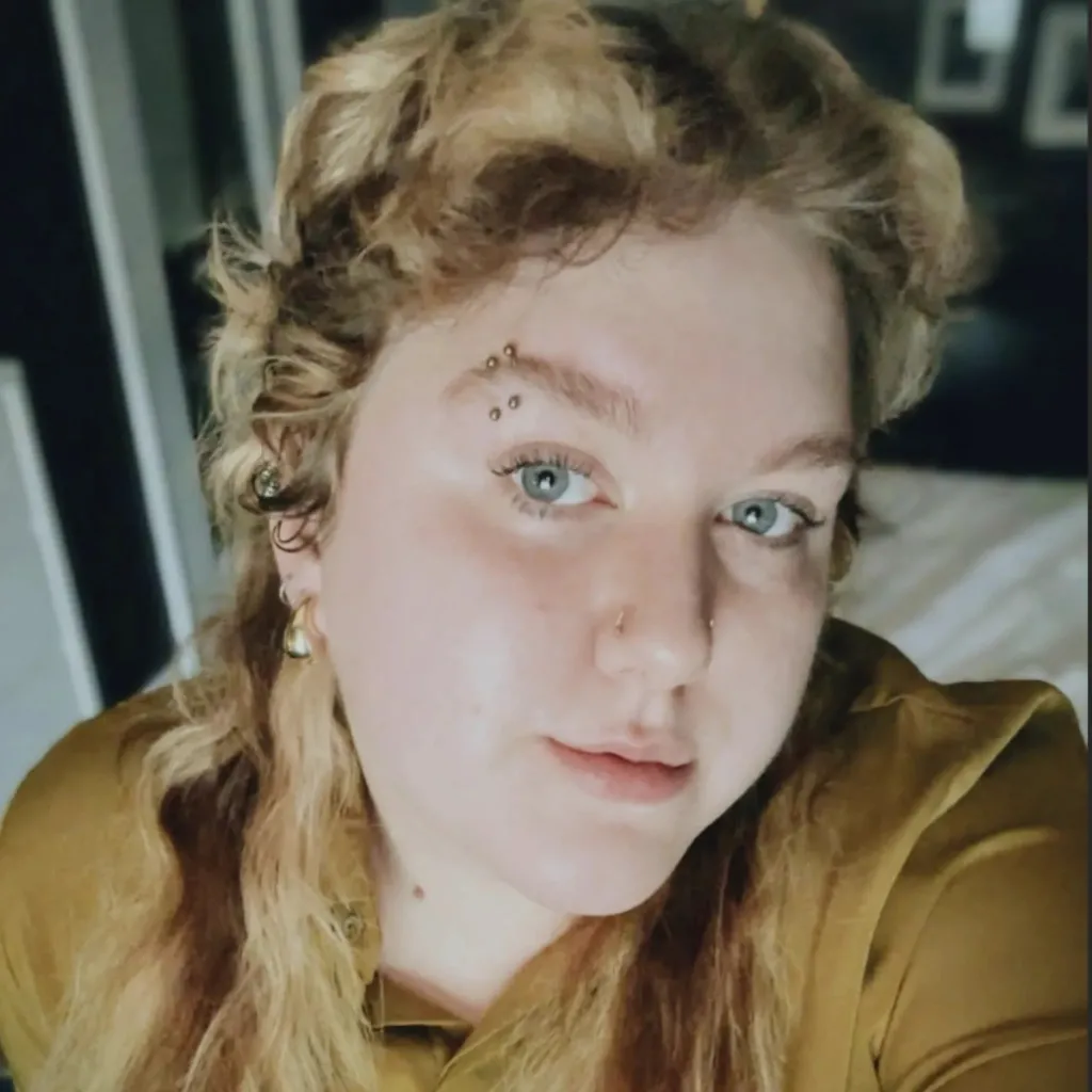 Headshot of Lived Experience (LEx) Leader Brina Williams wearing a gold shirt with curly blond hair and gold earrings in an indoor setting.